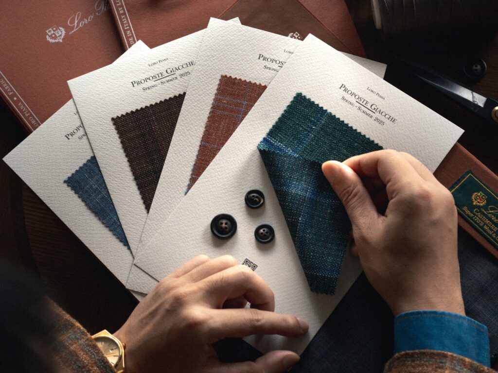A top-down view shows a person's hands, possibly a man's, with a gold watch on the left wrist, interacting with several fabric swatch books. The hands are holding up a teal-blue checkered fabric swatch from a book titled 'Loro Piana Proposte Giacche Spring-Summer 2025'. Other fabric swatches in shades of brown, blue, and a darker brown plaid are fanned out beneath it. Three dark, possibly horn or corozo, suit buttons are placed near the center. The background features other fabric books or textiles, including one with a 'Loro Piana' label visible.