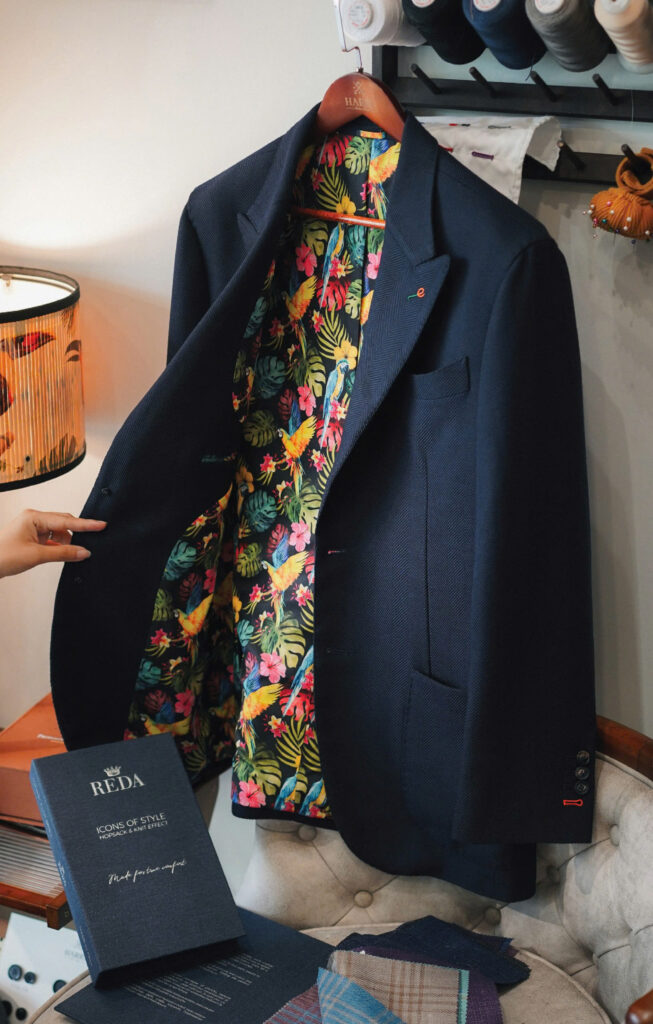 Navy bespoke jacket made from REDA wool hopsack fabric, featuring a vibrant floral lining, displayed on a wooden hanger in a tailor studio.