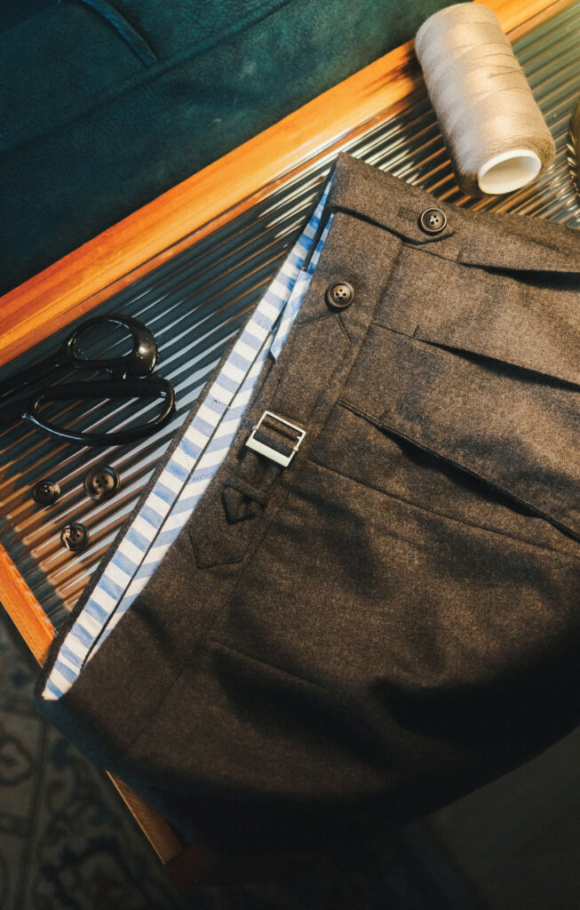 Close-up of bespoke wool trousers with sidetab and striped lining, displayed alongside tailoring tools on a wooden surface.