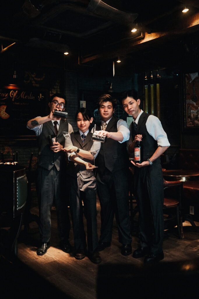 Bartenders at Merlin Cocktail & Eatery in Ho Chi Minh City wearing bespoke uniforms, posing behind the bar with shakers and a bottle of Campari — highlighting bespoke uniform tailoring.